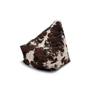 May include: A beanbag chair with a cowhide pattern. The chair is triangular in shape, with a white base and a brown and white spotted pattern on the top. The beanbag is filled and has a soft appearance.
