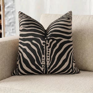 May include: Decorative throw pillow with a zebra stripe pattern in black and tan. The pillow is square and rests on a beige sofa. The zebra print is a bold, graphic design, adding a touch of safari style to the room.
