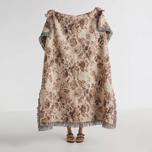 Puede incluir: Una manta grande y rectangular con un estampado floral en tonos marrones y beige. La manta tiene un ribete de flecos en los bordes. La manta se sostiene, mostrando el diseño y la textura completos.