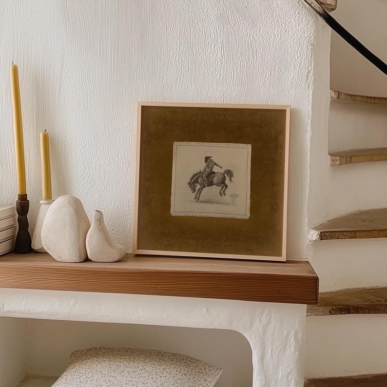 May include: A framed equestrian print with a brown mat and light wood frame. The print depicts a rider on horseback. Two yellow candles and white decorative objects sit on a wooden shelf. The background is a white wall.