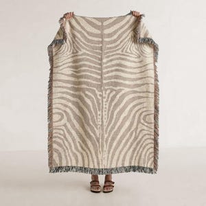 May include: A rectangular woven throw blanket featuring a beige and cream zebra stripe pattern. The blanket has a textured surface with a fringe border along all four edges, displaying a consistent animal-inspired design throughout.