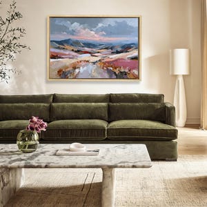 May include: An abstract landscape painting in a wooden frame, featuring a valley with pink, beige, and blue hues. The painting hangs above a green velvet sofa, with a marble coffee table in front.
