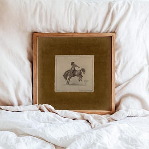 May include: A framed pencil sketch of a cowboy riding a bucking bronco. The artwork is set against a dark olive green background within a wooden frame. The image is displayed on a white bedspread.