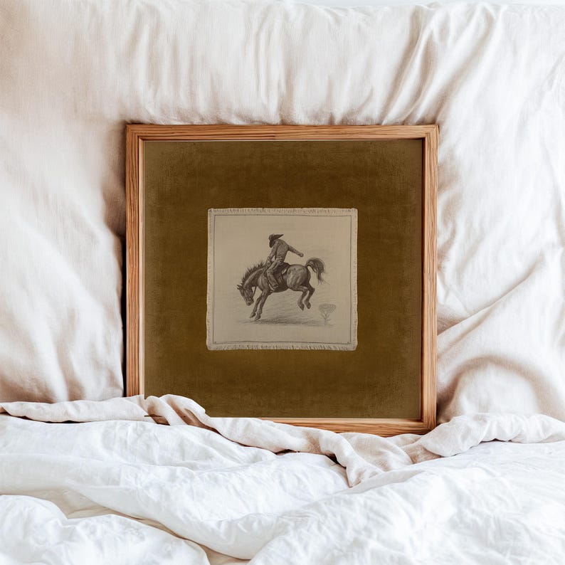 May include: A framed pencil sketch of a cowboy riding a bucking bronco. The artwork is set against a dark olive green background within a wooden frame. The image is displayed on a white bedspread.
