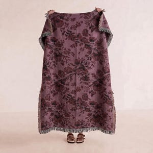May include: A rectangular woven throw blanket in a deep mauve color featuring a dark floral pattern of roses and leaves. The blanket has a textured finish and is bordered by short, frayed fringe edges along the sides and the bottom hem.