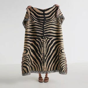 Può includere: Una grande coperta rettangolare con un motivo a strisce di zebra in nero e beige. La coperta ha un bordo con frange. Il design ricorda la pelle di una zebra. La coperta è tenuta da una persona.
