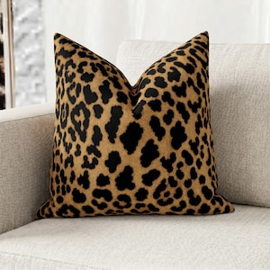 Pode incluir: Almofada quadrada com estampa de leopardo. A almofada apresenta um fundo bege com manchas pretas, imitando o padrão de um leopardo. Está colocada em um sofá de cor clara, adicionando um toque decorativo ao espaço.