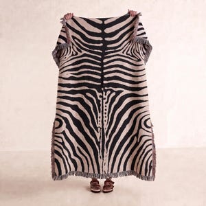 Puede incluir: Una manta rectangular tejida con un patrón de rayas de cebra de alto contraste en negro y beige. El tejido presenta un diseño simétrico con una columna central y detalles de flecos decorativos en los bordes superior e inferior.