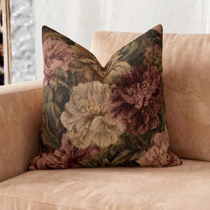 May include: A square throw pillow featuring a dark, moody floral pattern with large, detailed peony blossoms in shades of deep burgundy, dusty rose, and cream, set against a background of dark green foliage and shadows on a soft fabric surface.