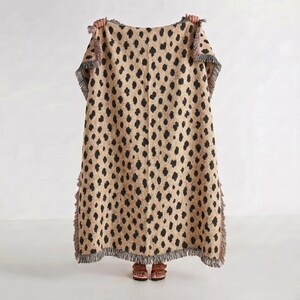 May include: A rectangular woven throw blanket featuring a tan base color with a repeating pattern of irregular black spots. The blanket has a textured appearance and is finished with short, frayed fringe detailing along all four edges of the fabric.