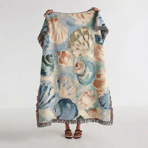 May include: A rectangular throw blanket featuring a seashell pattern in shades of blue, beige, and brown. The blanket has a fringed edge and is held up by a person. The design includes various seashell illustrations.
