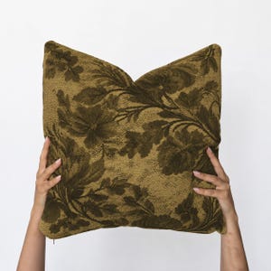 May include: A square throw cushion featuring a textured, woven fabric in shades of olive green and dark brown. The surface displays a dense, repeating floral pattern with large leaves and blossoms that cover the entire front of the cushion.