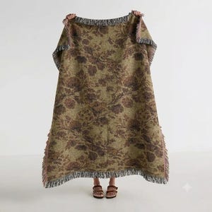Pode incluir: Uma manta retangular tecida com um padrão floral castanho escuro sobre um fundo verde-azeitona suave. A manta tem uma aparência texturizada com bordas de franjas desfiadas e multicoloridas nas extremidades superior e inferior.