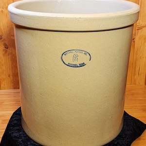 May include: A large, beige stoneware crock with a blue and white oval label that reads "Marshall Pottery, Inc. 6 Marshall, Texas".