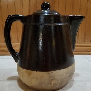 May include: A brown and tan glazed ceramic teapot with a lid. The teapot has a wide base and a narrow spout.