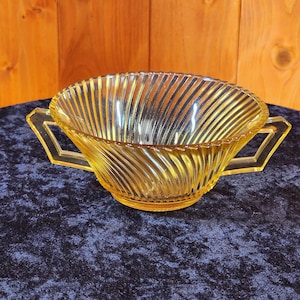 May include: A vintage yellow glass bowl with a ribbed design and two angular handles. The bowl has a curved shape and sits on a dark blue surface. The glass has a slight amber tint, and the handles are a darker shade of yellow.