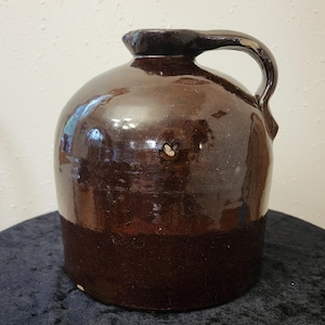 May include: A dark brown glazed stoneware jug with a rounded body and a single side handle. The surface features a glossy finish with visible variations in the brown tone and small areas of wear along the handle and body of the vessel.