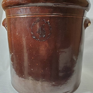 May include: A brown glazed stoneware crock with a circular embossed design of a sailboat. The crock has two handles on the sides.
