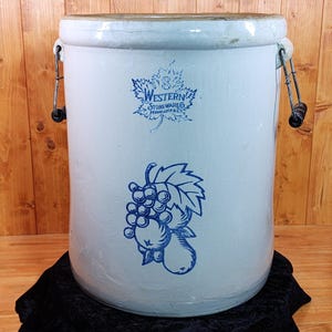 May include: A large, cylindrical, off-white stoneware crock featuring a blue maple leaf logo with the text '8 Western Stoneware Co. Monmouth Ill.' and a blue graphic of a cluster of grapes and fruit on the front, with two metal side handles.