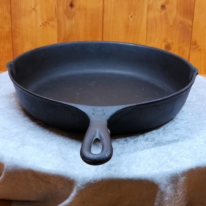 May include: A black cast iron skillet with a long handle and a rounded cooking surface. The skillet is sitting on a white surface. The handle has a hole for hanging. The skillet is a classic kitchen tool.