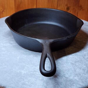 May include: A black cast iron skillet with a single handle featuring a hole for hanging. The skillet has a wide, shallow cooking surface and a slightly raised rim. The handle has a small "S" marking.