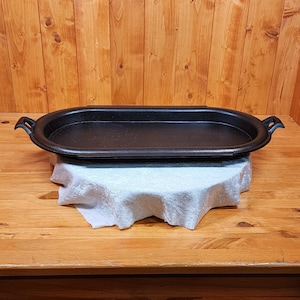 May include: A black, oval-shaped cast iron griddle with handles. The griddle is sitting on a white cloth, and the background is a wooden surface. This kitchenware is designed for cooking.