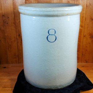 May include: A large, off-white stoneware crock with a blue number 8 painted on the front. The crock has a wide opening and a rounded rim. It is displayed on a black surface against a wooden background.