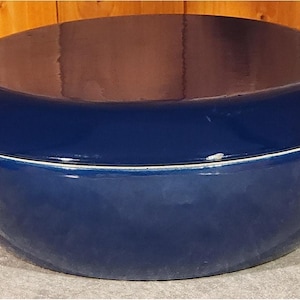 May include: A deep blue, oval-shaped Dutch oven with a matching lid. The pot has two handles and a white accent line around the rim. The pot sits on a light-colored surface.