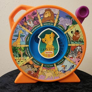 May include: A round orange plastic See 'n Say toy featuring The Lion King characters. The center dial displays Simba with the text The Lion King. Surrounding sections show Mufasa, an elephant, Pumbaa, a zebra, Zazu, Nala, Ed, Timon, a giraffe, and Rafiki.