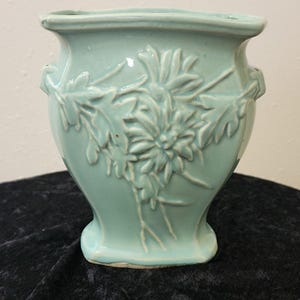May include: A light mint green ceramic vase featuring a raised relief design of chrysanthemum flowers and leaves on the front. The vessel has a flared rim, a tapered body, and small decorative handles on each side with a smooth glossy finish.