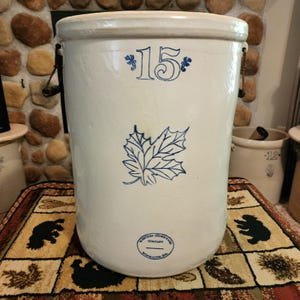 May include: A large off-white stoneware crock featuring a blue number 15 at the top, a blue maple leaf illustration in the center, and a blue Western Stoneware Company Monmouth, Ill. stamp at the bottom, with two metal side handles.