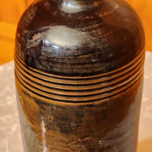 May include: A dark brown ceramic bottle with horizontal, ridged detailing. The bottle has a glossy finish with areas of lighter brown and tan. The bottle's shape is rounded at the top and tapers towards the base, suggesting a vintage or antique style.