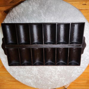 May include: A dark brown cast iron muffin pan with twelve individual compartments. The pan has a rectangular shape with rounded edges and a handle on each end. The pan is sitting on a white, textured surface.
