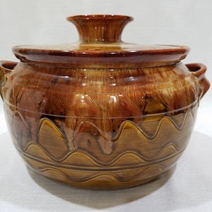 Puede incluir: Una cazuela de cerámica cubierta con un rico esmalte marrón. El plato presenta un patrón ondulado decorativo alrededor de la base y dos asas. La tapa tiene un pomo para facilitar el manejo. Un artículo de menaje rústico.