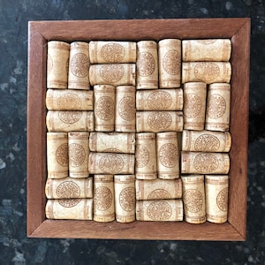May include: A wooden frame with a cork trivet inside. The trivet is made of a grid of wine corks, each with a circular design.