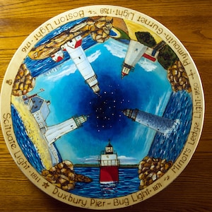 May include: A wooden round plate with a painted design of lighthouses and their names and dates. The lighthouses are painted in various colors, including red, white, and blue. The plate is decorated with a blue and white ocean scene.
