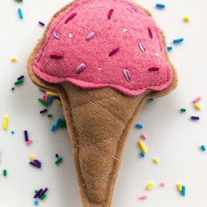 Puede incluir: Un cono de helado de fieltro rosa con chispas blancas y moradas. El cono es marr&oacute;n y tiene una textura de waffle. El helado est&aacute; rodeado de chispas de colores.