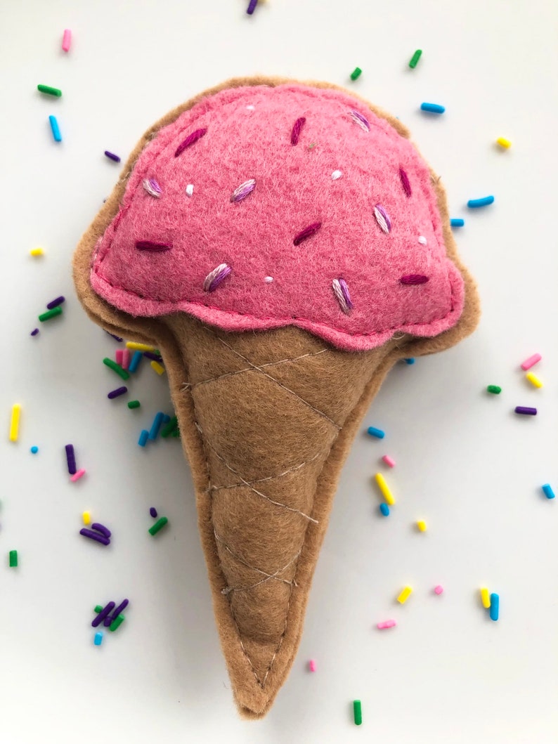 Puede incluir: Un cono de helado de fieltro rosa con chispas blancas y moradas. El cono es marr&oacute;n y tiene una textura de waffle. El helado est&aacute; rodeado de chispas de colores.