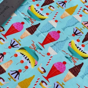May include: Pale blue fabric featuring a repeating pattern of ice cream cones, sundaes, and banana splits. The ice cream cones have pink, brown, and yellow cones. The sundaes and banana splits have red cherries. A stainless steel ruler is in the upper left corner.