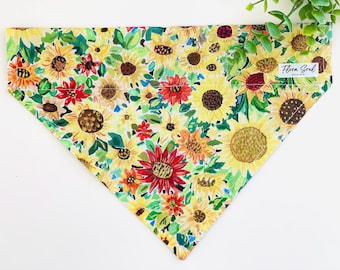 May include: A yellow, green, red, and brown dog bandana with a sunflower print. The bandana has a white label with the text "Flora Soul Design".