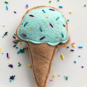 Puede incluir: Un cono de helado azul claro con chispas, hecho de fieltro. El cono es marr&oacute;n y el helado es azul claro con chispas de colores.
