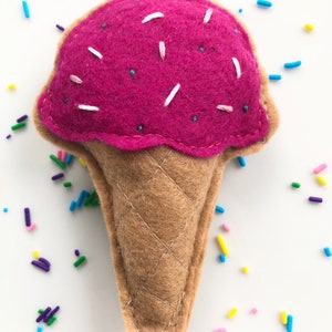Puede incluir: Un cono de helado de fieltro rosa con chispas blancas, sobre un fondo blanco.