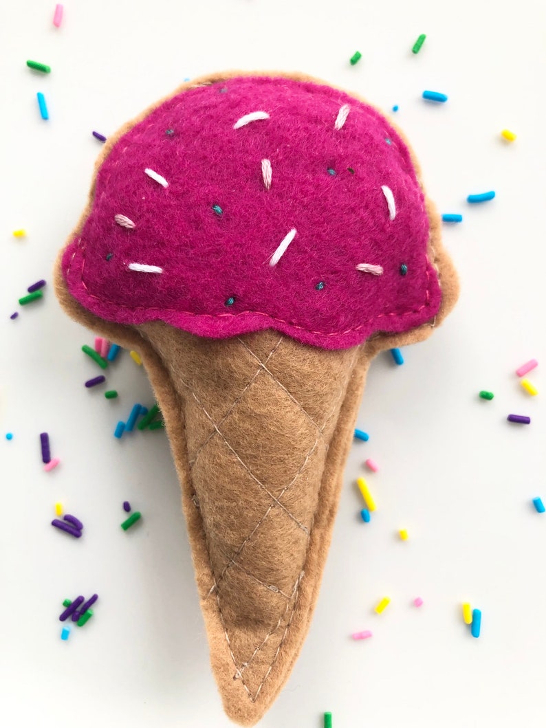 Puede incluir: Un cono de helado de fieltro rosa con chispas blancas, sobre un fondo blanco.