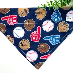 Op de afbeelding: Een donkerblauwe hondenbandana met een baseballthema. De bandana heeft baseballs, handschoenen en nummer één schuimvingers in rood, wit en blauw. De bandana heeft de tekst "Flora Soul Design" in de rechterbovenhoek.