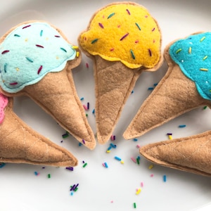May include: Five felt ice cream cones with different coloured ice cream and sprinkles. The ice cream cones are light brown and the ice cream is pink, yellow, blue, and light blue.