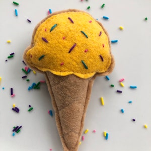 Puede incluir: Un cono de helado de fieltro amarillo con chispas de colores. El cono es marr&oacute;n y tiene una superficie texturizada.