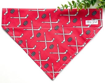 May include: Red dog bandana with a white hockey stick and puck pattern. The bandana has a white label with the text "Flora Soul Design".