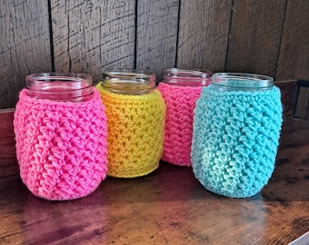 May include: Four glass jars with crocheted covers in pink, yellow, pink, and blue. The jars are sitting on a wooden surface.