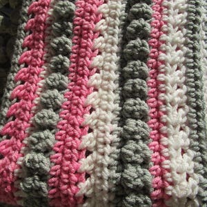 May include: A striped crocheted blanket with pink, white, and gray yarn. The blanket is made with a textured stitch that creates a bumpy surface.