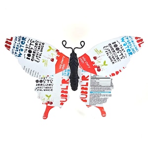 May include: A whimsical butterfly sculpture crafted from repurposed beverage packaging. The wings feature text and cherry illustrations, with the words "ANTIOXIDANT SPARKLING WATER" and "BOOSTS ENERGIZES RESTORES BALANCE". The body is black.